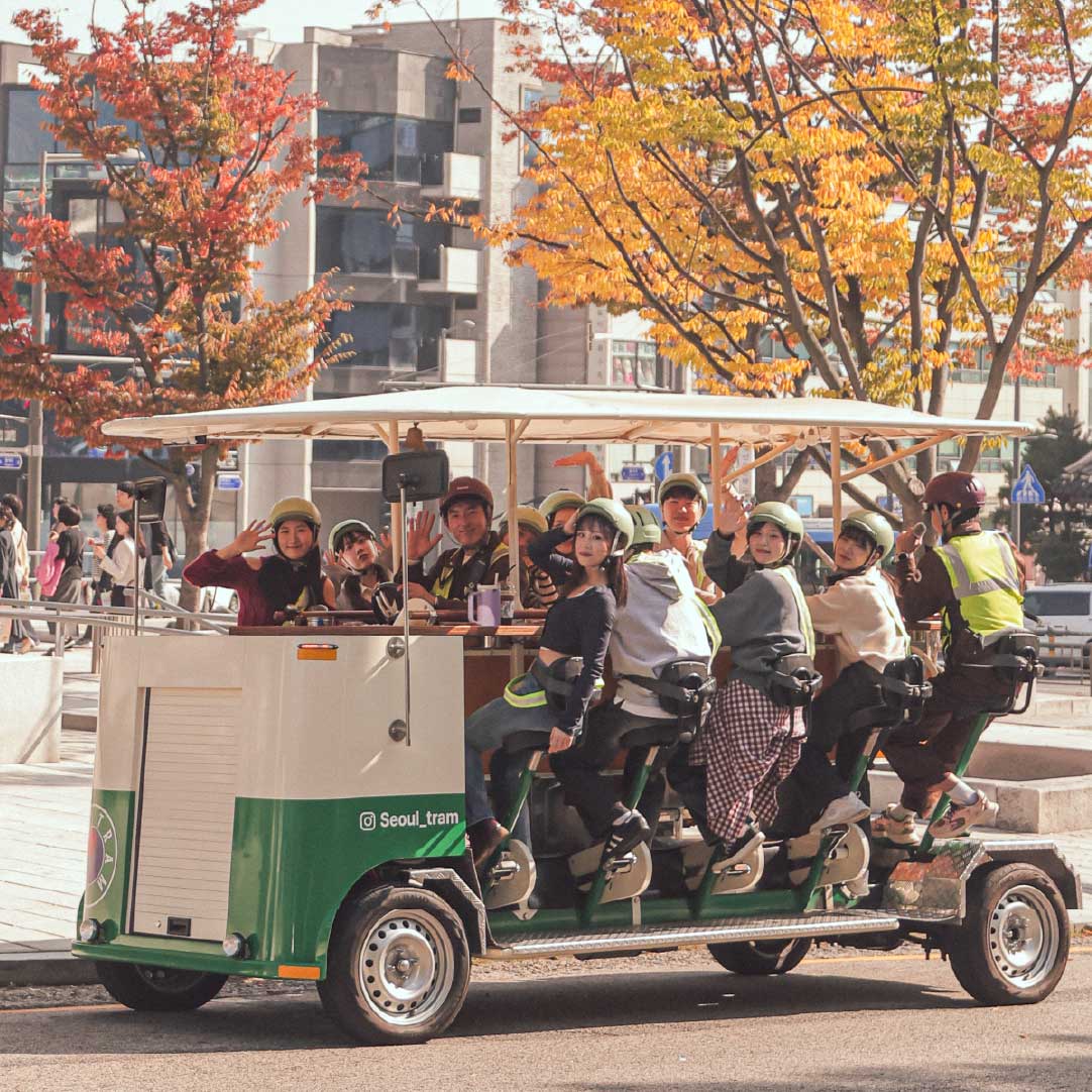 A group of tourists smiling and waving while riding the Seoul Tram in front of vibrant fall trees. 一群游客在缤纷秋叶背景下乘坐首尔电车，微笑挥手。