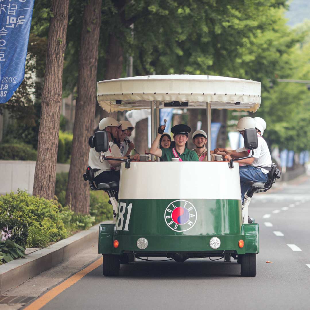 Group of people smiling and pedaling on the Seoul Tram through tree-lined streets in summer 一群人在夏季绿荫街道上骑行首尔电车，笑容满面